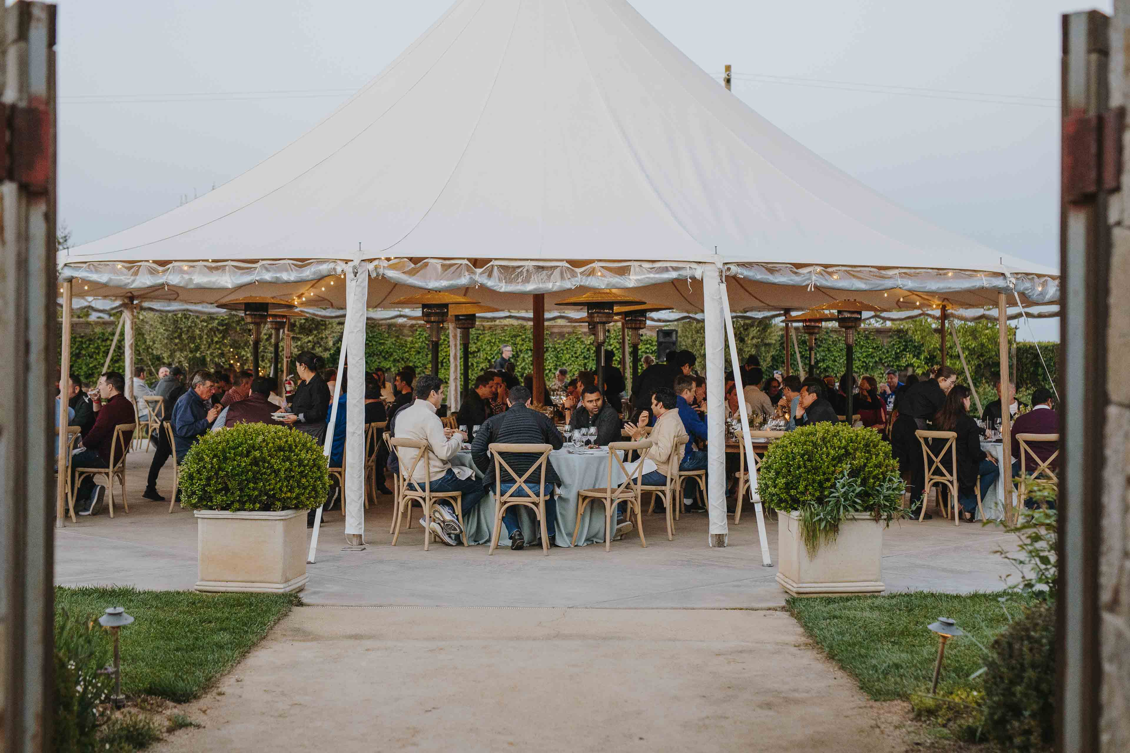 People Sitting At Tables Under A Large White Tent