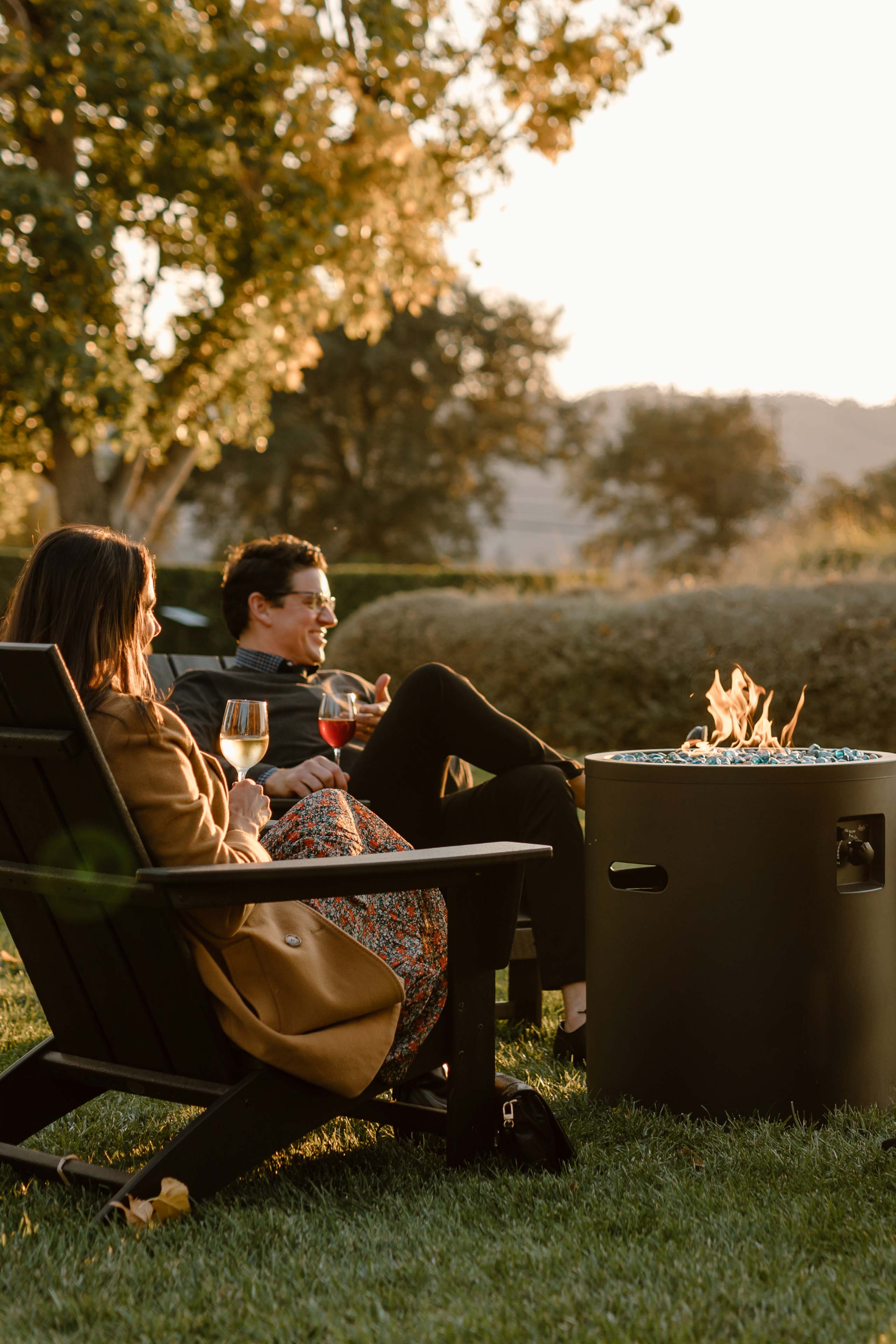 Two People Reclining In Chairs Near A Fire Pit Holding Glasses Of Wine